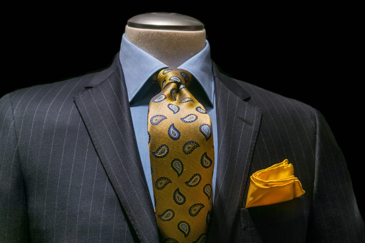 Close-up of a gray striped jacket with blue shirt, patterned golden yellow tie and handkerchief on black background